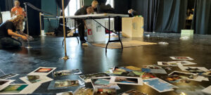 bannerbillede som viser sider og fotografier spredt på et gulv, mens børn arbejder i baggrunden. Billedet er fra en workshop for elever på Teatret Zeppelin.
